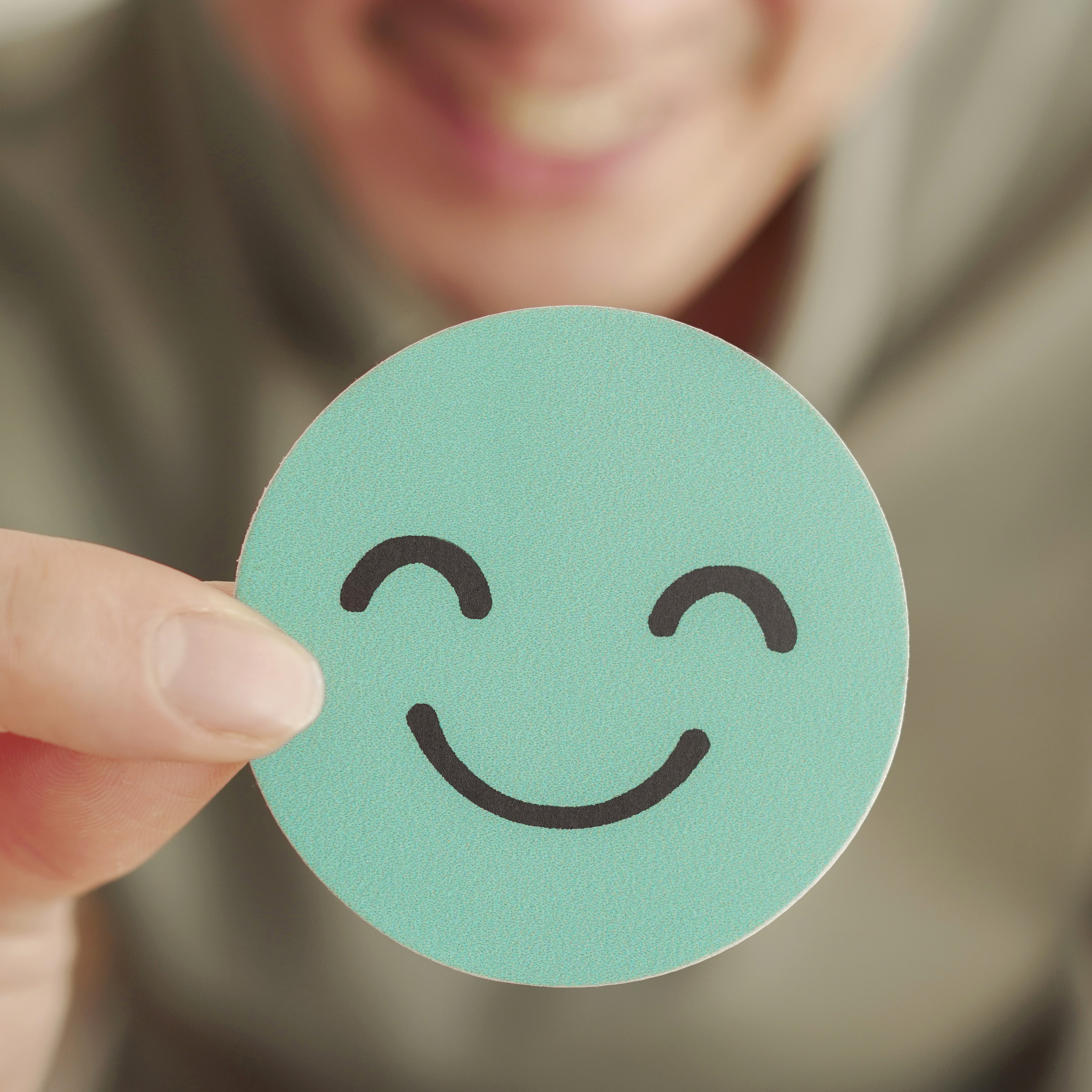 Person hält ein Stück Papier mit einem lächelnden, grünen Gesicht darauf, in die Kamera